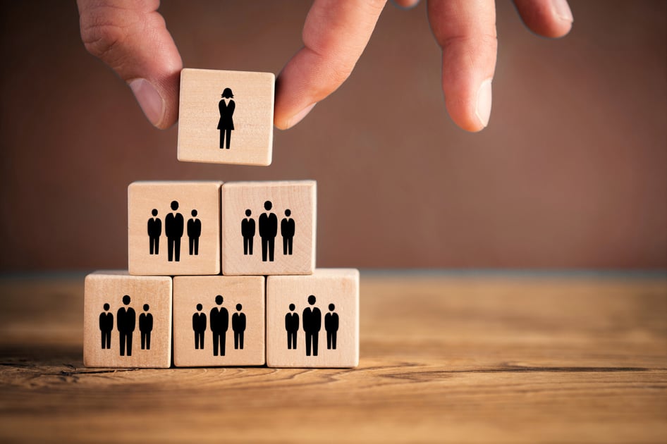 Hand plaatst houten blokje met een pictogram van een persoon boven op een piramide van andere blokjes, symbool voor verhoogde verantwoordelijkheid van leidinggevenden.