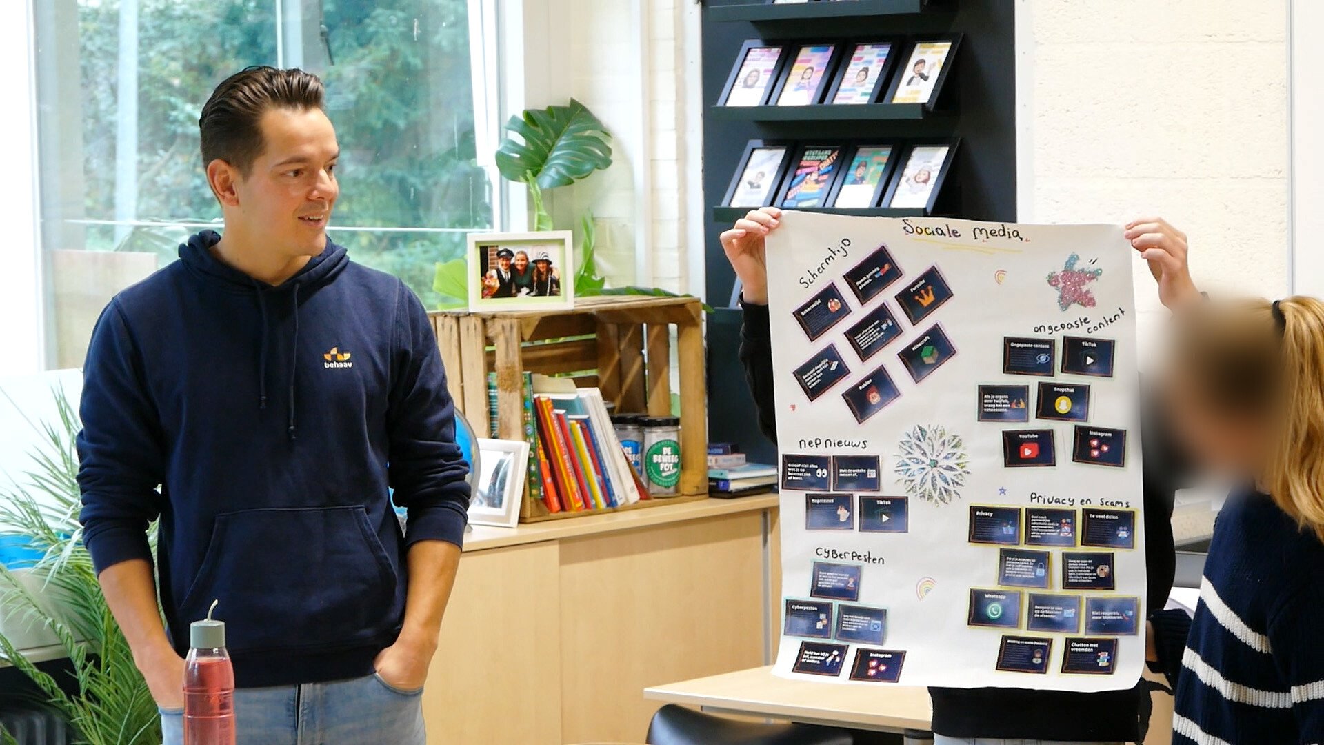 Een man in een donkerblauwe hoodie staat in een klaslokaal en kijkt naar twee studenten die een groot posterbord presenteren over omgaan met sociale media.